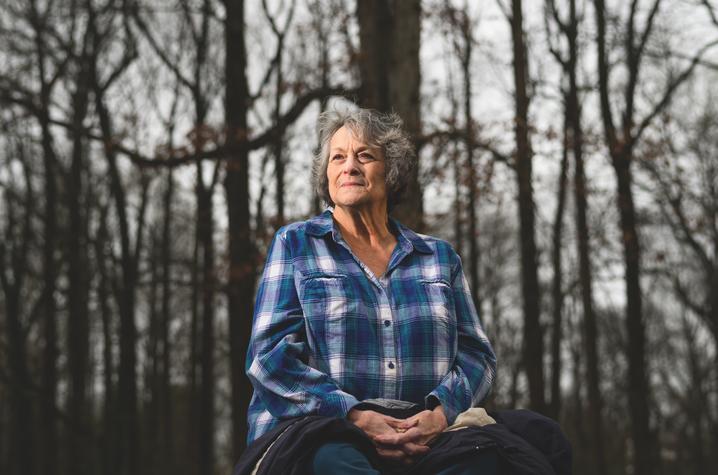 Woman in a plaid shirt sitting in front of trees, looking off. 