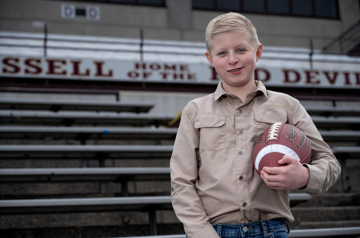 Boy standing in front of bleachers in a tan shirt, holding a football. 