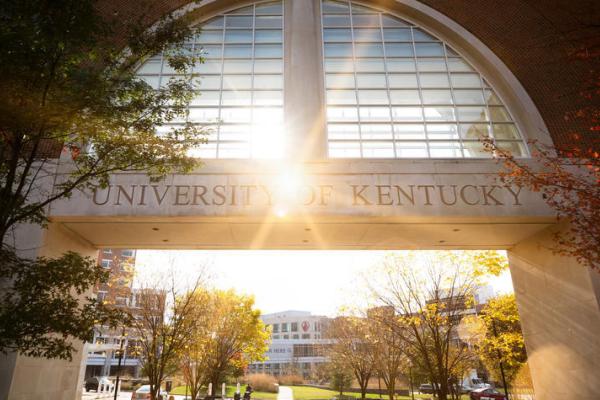 Picture of the sun shining through a glass structure with the words "University of Kentucky" carved into stone."