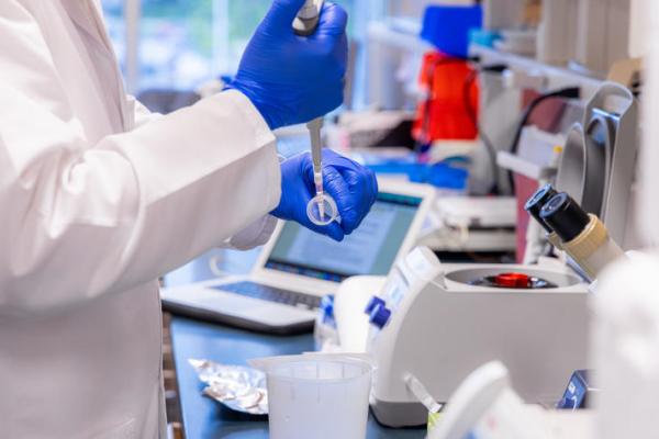 Person in white coat and blue gloves measuring out a liquid. 