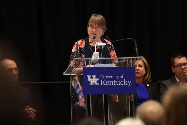 Woman in floral blouse speaking behind a glass podium. 