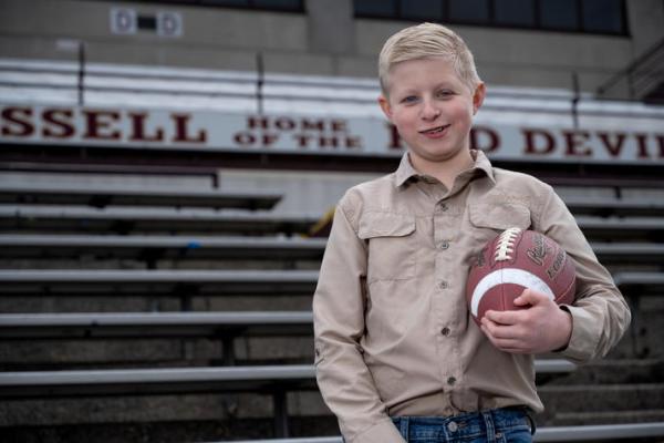 Boy standing in front of bleachers in a tan shirt, holding a football. 