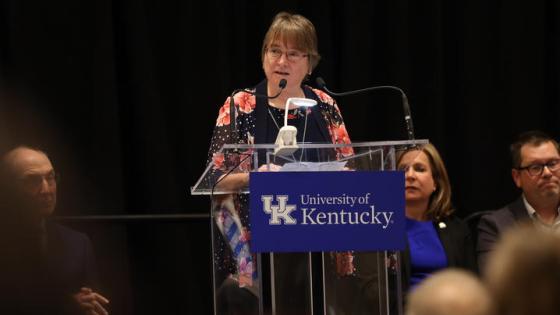 Woman in floral blouse speaking behind a glass podium. 