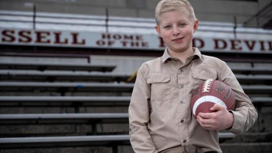 Boy standing in front of bleachers in a tan shirt, holding a football. 